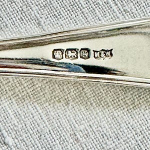 May include: Close-up of a silver spoon with hallmarks. The spoon has a reflective surface and is set against a textured, light gray background. The hallmarks include a crown, a lion, and other symbols, along with the initials "M&W".