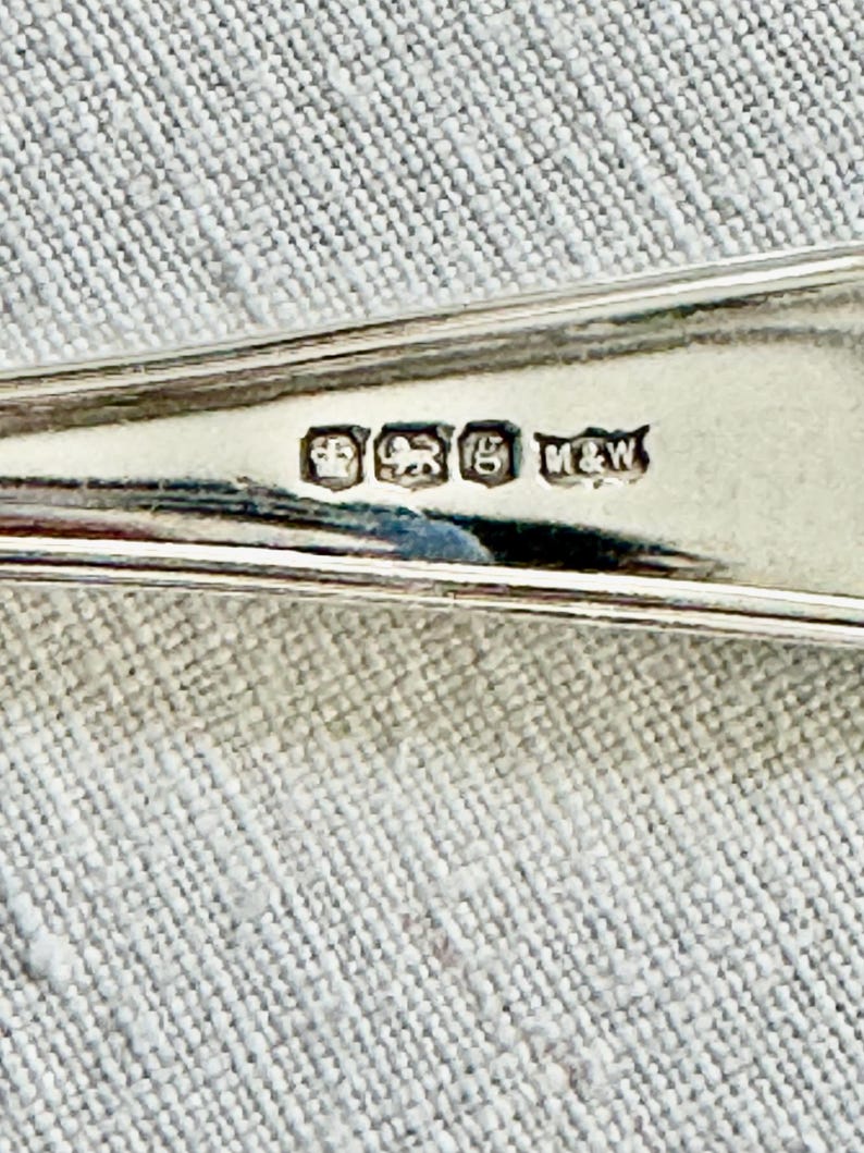 May include: Close-up of a silver spoon with hallmarks. The spoon has a reflective surface and is set against a textured, light gray background. The hallmarks include a crown, a lion, and other symbols, along with the initials "M&W".