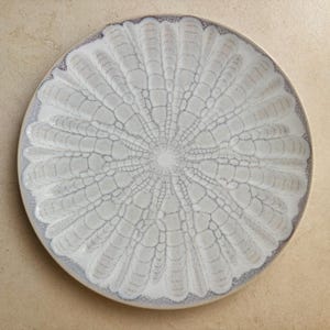 May include: A round ceramic plate featuring a radial pattern of elongated, petal-like shapes in shades of off-white and cream. The design is defined by dark, intricate crackle-like lines that radiate from the centre towards the outer edge.