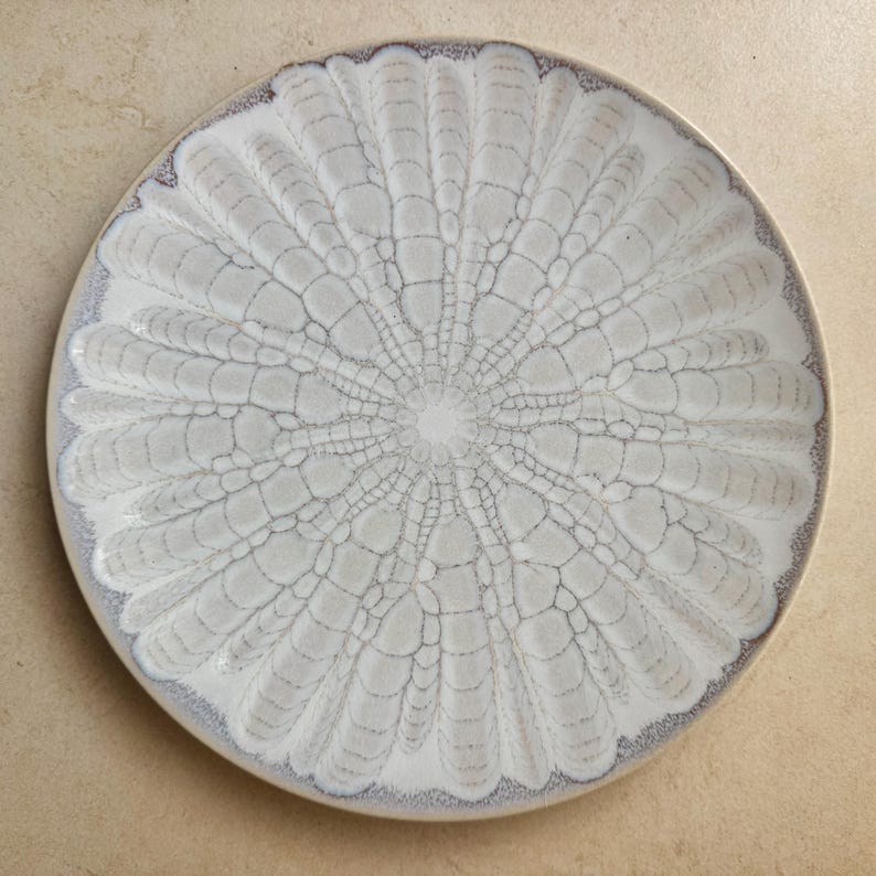 May include: A round ceramic plate featuring a radial pattern of elongated, petal-like shapes in shades of off-white and cream. The design is defined by dark, intricate crackle-like lines that radiate from the centre towards the outer edge.