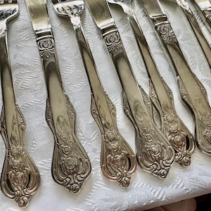 May include: A collection of silver-colored forks and knives with ornate floral detailing on the handles. The silverware is arranged on a white patterned tablecloth, showcasing a classic and elegant design.