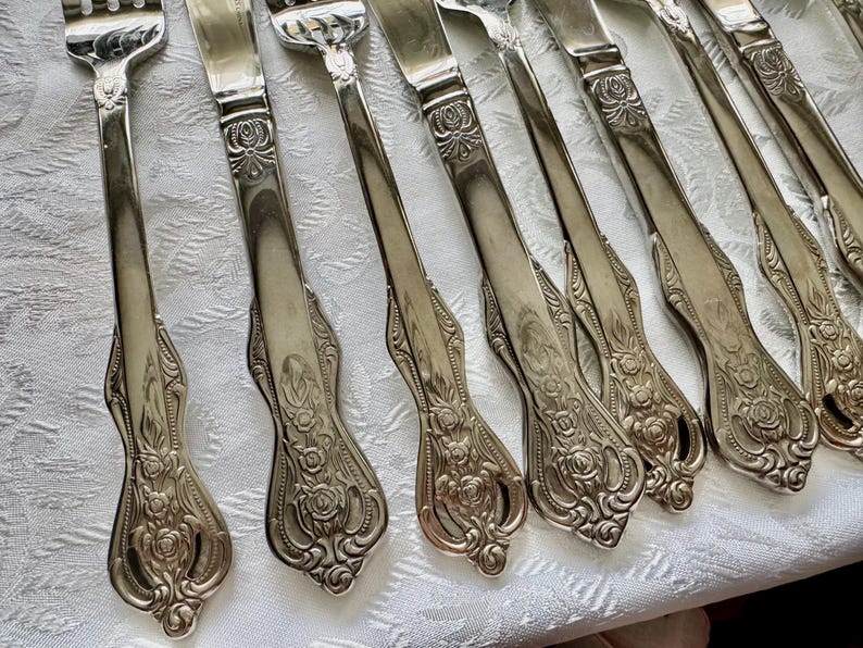 May include: A collection of silver-colored forks and knives with ornate floral detailing on the handles. The silverware is arranged on a white patterned tablecloth, showcasing a classic and elegant design.