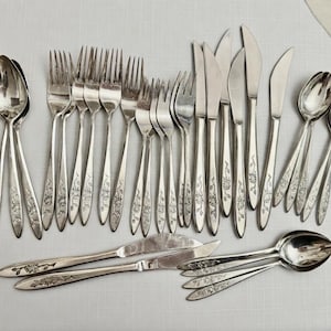 May include: A set of vintage silverware, comprising spoons, forks, and knives, displayed on a light surface. The cutlery features a floral pattern on the handles. The set includes different sizes of spoons, forks, and knives.
