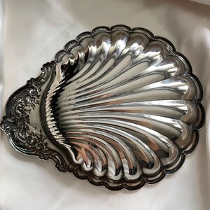 K&ouml;nnte beinhalten: Eine polierte, silberfarbene Metallschale in Form einer Jakobsmuschel. Die Schale weist ein tief geriffeltes, strahlenf&ouml;rmiges Design &uuml;ber dem Hauptbecken sowie ein verziertes, gepr&auml;gtes Blumen- und Rankenmuster am griffartigen Ansatz auf.