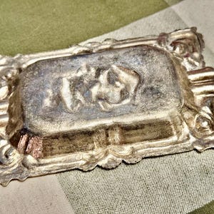 May include: A rectangular metallic ashtray with a tarnished silver finish. The center features a raised relief design of a reclining figure. The border is decorated with ornate, scrolling patterns and includes two integrated cigarette rests.