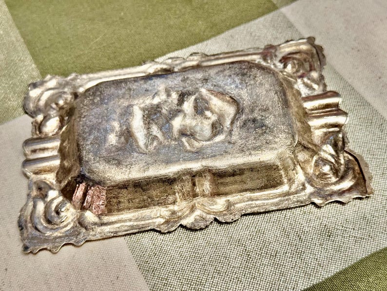 May include: A rectangular metallic ashtray with a tarnished silver finish. The center features a raised relief design of a reclining figure. The border is decorated with ornate, scrolling patterns and includes two integrated cigarette rests.