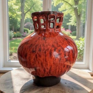 May include: A large, glossy, red-orange ceramic vase with a bulbous body and a dark brown, openwork top. The vase has a dark brown base and sits on a wooden surface, with a bright, natural background visible through a window.