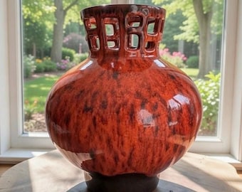 Puede incluir: Un jarrón grande de cerámica rojo-naranja brillante, con un cuerpo bulboso y una parte superior calada de color marrón oscuro. El jarrón tiene una base marrón oscuro y se asienta sobre una superficie de madera, con un fondo natural brillante visible a través de una ventana.