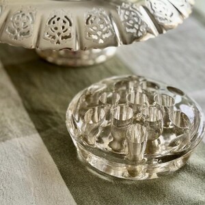 May include: A clear glass flower frog with multiple holes sits on a green and white striped surface. A silver-colored, decorative pedestal dish is in the background. The flower frog is round and has a clear, transparent appearance.