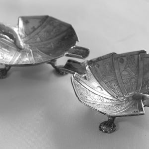 K&ouml;nnte beinhalten: Zwei silberfarbene, dekorative Aschenbecher. Jeder Aschenbecher hat eine f&auml;cherartige Form mit aufwendigen ge&auml;tzten Designs und ruht auf drei kleinen, klauenartigen F&uuml;&szlig;en. Die Aschenbecher haben eine kleine, gebogene Ablage f&uuml;r eine Zigarette.