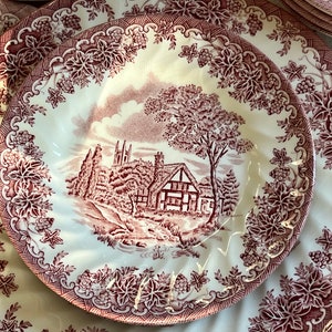 May include: A set of vintage ceramic plates with a cottagecore design. The plates feature a red and white floral pattern with a cottage scene in the center. The cottage is surrounded by trees and a stream.