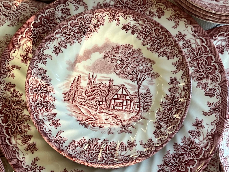 May include: A set of vintage ceramic plates with a cottagecore design. The plates feature a red and white floral pattern with a cottage scene in the center. The cottage is surrounded by trees and a stream.