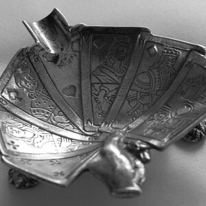 K&ouml;nnte beinhalten: Ein silberfarbener Spielkartenhalter in Form einer Hand, die Karten auff&auml;chert. Die Karten zeigen detaillierte Illustrationen von K&ouml;nigen, K&ouml;niginnen und Spielkartensymbolen. Der Halter steht auf kleinen, dekorativen F&uuml;&szlig;en.