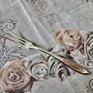 May include: A silver, two-tined fork with an ornate handle rests on a floral patterned tablecloth. The fork's tines are slender and pointed. The tablecloth features a repeating pattern of roses in shades of beige and gray.