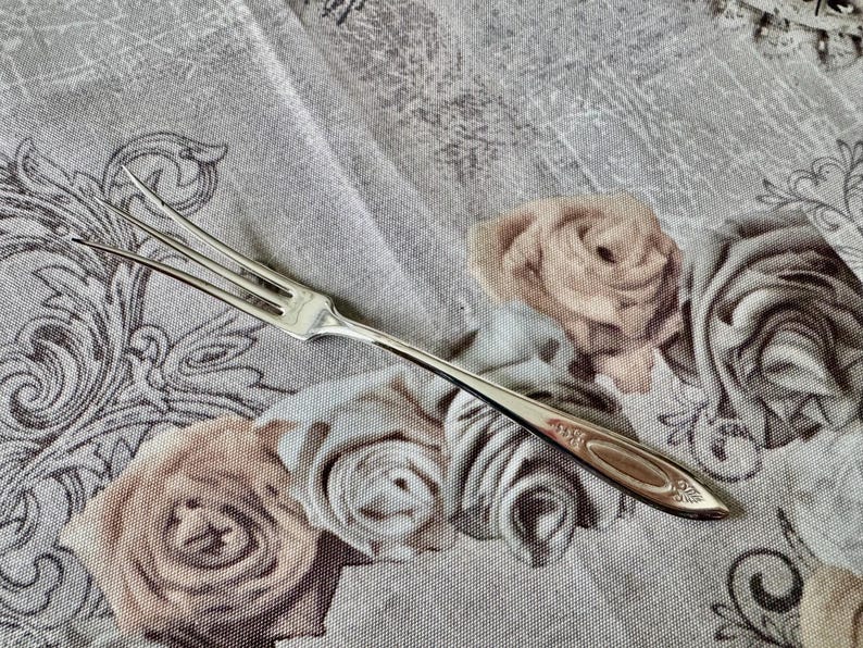 May include: A silver, two-tined fork with an ornate handle rests on a floral patterned tablecloth. The fork's tines are slender and pointed. The tablecloth features a repeating pattern of roses in shades of beige and gray.