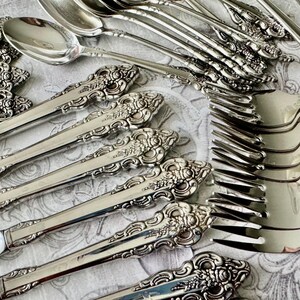 May include: Close-up of a collection of ornate silver silverware, including forks, spoons, and knives. The silverware has detailed floral and scrollwork patterns on the handles. The arrangement suggests a formal dining setting.