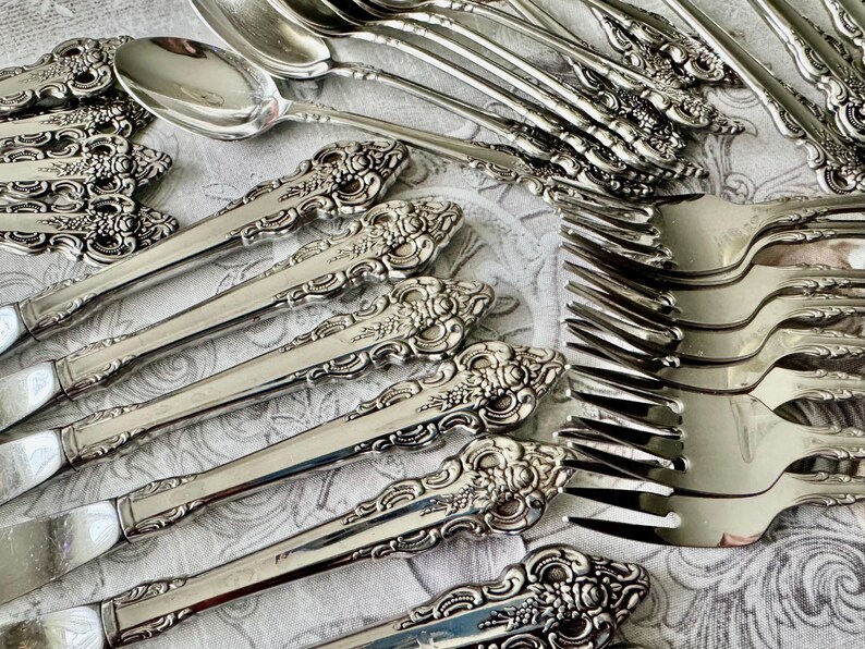 May include: Close-up of a collection of ornate silver silverware, including forks, spoons, and knives. The silverware has detailed floral and scrollwork patterns on the handles. The arrangement suggests a formal dining setting.