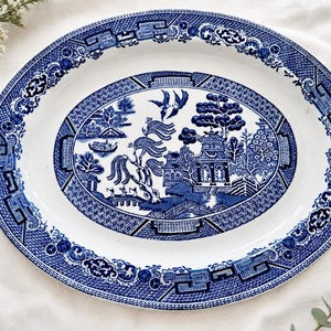 May include: An oval ceramic serving platter featuring a classic blue and white willow pattern. The design includes detailed illustrations of a pagoda, a bridge, willow trees, and two birds in flight, surrounded by a decorative geometric border.