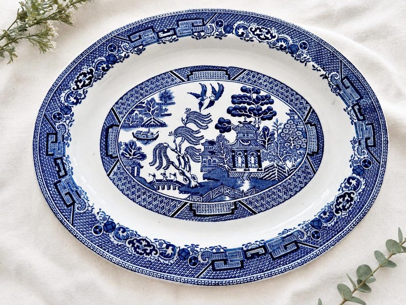 May include: An oval ceramic serving platter featuring a classic blue and white willow pattern. The design includes detailed illustrations of a pagoda, a bridge, willow trees, and two birds in flight, surrounded by a decorative geometric border.