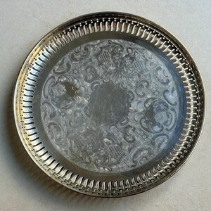 Puede incluir: Una bandeja redonda de color plateado con un patr&oacute;n floral grabado y ornamentado. La bandeja tiene un borde decorativo y estriado. La bandeja es probablemente de metal y es adecuada para servir alimentos o exhibir art&iacute;culos.