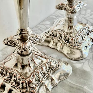 May include: Two silver-colored metal candle holders featuring square bases with ornate, raised leaf-like scrollwork patterns. The stems are smooth and tapered, rising from the center of the detailed, decorative square pedestal bases.