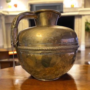 K&ouml;nnte beinhalten: Ein antiker Messingkrug mit rundem Korpus und einem einzelnen Henkel. Der Krug hat ein dekoratives Band um die Mitte.