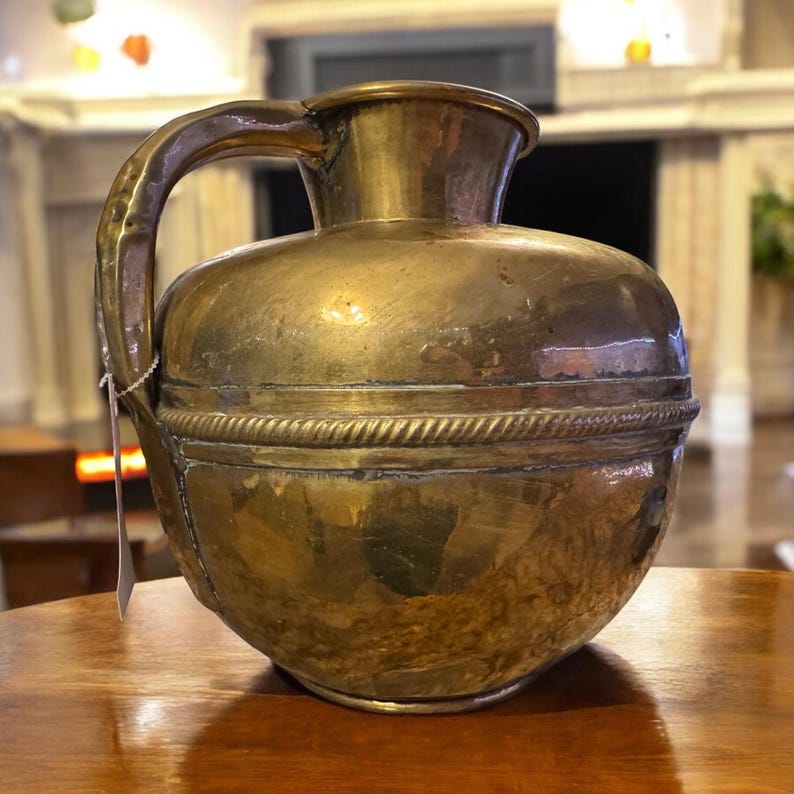 K&ouml;nnte beinhalten: Ein antiker Messingkrug mit rundem Korpus und einem einzelnen Henkel. Der Krug hat ein dekoratives Band um die Mitte.