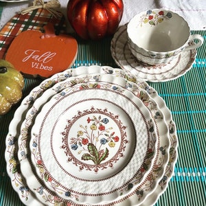 May include: Three white ceramic plates with a brown, floral pattern and scalloped edges. The plates are stacked on top of each other and are sitting on a green and blue bamboo mat. The plates have a woven pattern and feature a floral design with red, yellow, and blue flowers.