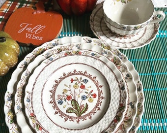 May include: Three white ceramic plates with a brown, floral pattern and scalloped edges. The plates are stacked on top of each other and are sitting on a green and blue bamboo mat. The plates have a woven pattern and feature a floral design with red, yellow, and blue flowers.