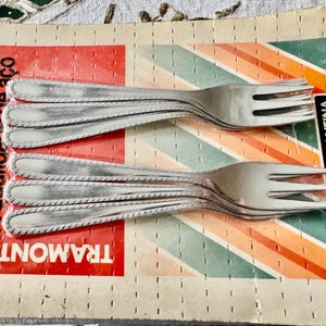 May include: A set of stainless steel forks on a red, orange, and green striped cardboard backing. The forks have a decorative edge and are labeled with the brand name "TRAMONTINA" and "Inox Stainless".