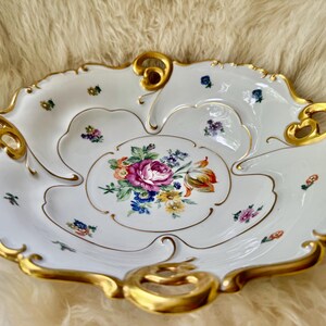 Puede incluir: Plato de porcelana blanca con borde festoneado y ribete dorado. El plato presenta un dise&ntilde;o floral en el centro con flores rosas, moradas y amarillas. Peque&ntilde;os acentos florales est&aacute;n dispersos.