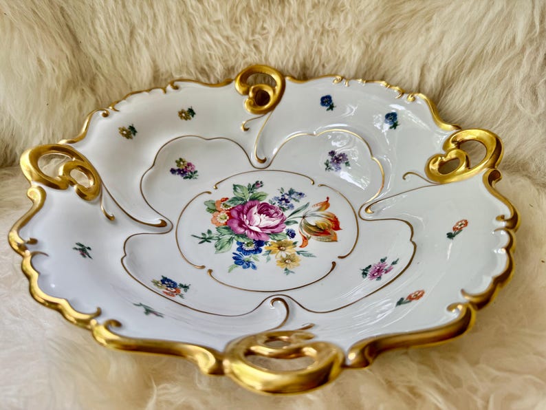 Puede incluir: Plato de porcelana blanca con borde festoneado y ribete dorado. El plato presenta un dise&ntilde;o floral en el centro con flores rosas, moradas y amarillas. Peque&ntilde;os acentos florales est&aacute;n dispersos.