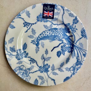 Puede incluir: Un plato redondo de cer&aacute;mica color crema con un patr&oacute;n azul de transferencia que muestra un ave de cola larga posada sobre ramas florecidas. Una etiqueta rectangular en la parte superior muestra el texto Queen's by Churchill y Made in England.