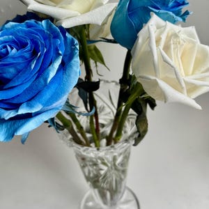 May include: A bouquet of fresh roses in shades of vibrant blue and crisp white, arranged in a clear, cut-glass crystal vase. The vase features a flared rim and a starburst pattern on its base, holding several long green stems with leaves.