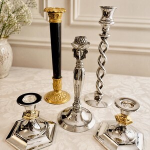 May include: A collection of five metal candlesticks. One features a tall black column with gold-toned ornate base and top. Two are short with hexagonal silver bases and gold-toned accents. One is a tall silver twisted stem, and one is a silver stem.