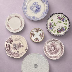 May include: A collection of seven ceramic plates and saucers featuring diverse designs, including blue and white floral patterns, purple landscape scenes, hand-painted fruit motifs, and a white plate with a textured, radiating petal pattern.