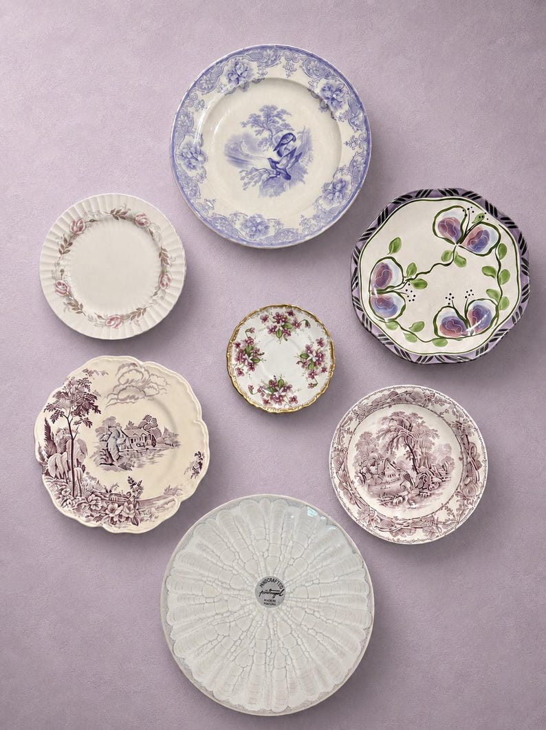 May include: A collection of seven ceramic plates and saucers featuring diverse designs, including blue and white floral patterns, purple landscape scenes, hand-painted fruit motifs, and a white plate with a textured, radiating petal pattern.