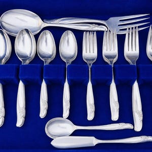 May include: A set of silver-toned flatware arranged in a blue velvet-lined case. The collection includes four spoons, four forks, one large serving spoon, one large serving fork, and one butter knife, all featuring a smooth, curved handle design.