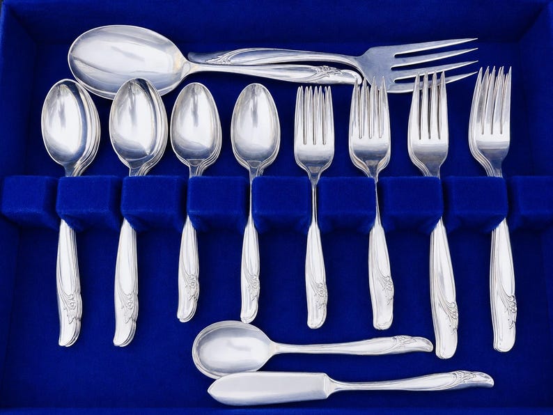 May include: A set of silver-toned flatware arranged in a blue velvet-lined case. The collection includes four spoons, four forks, one large serving spoon, one large serving fork, and one butter knife, all featuring a smooth, curved handle design.