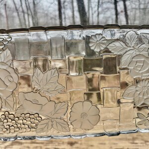 May include: A rectangular clear glass serving tray with a woven basket design. The tray is decorated with embossed floral and grape clusters. The edges are slightly scalloped. The tray is being held by a person.