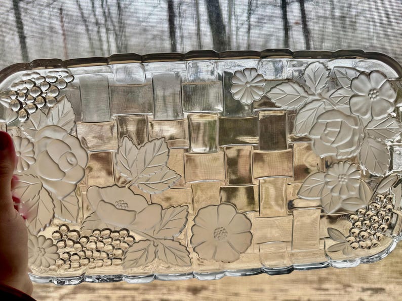 May include: A rectangular clear glass serving tray with a woven basket design. The tray is decorated with embossed floral and grape clusters. The edges are slightly scalloped. The tray is being held by a person.