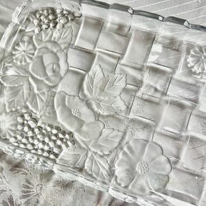 May include: A clear, rectangular glass serving tray with a woven basket design. The tray features embossed floral and leaf patterns, including roses, daisies, and clusters of berries. The edges of the tray are scalloped.