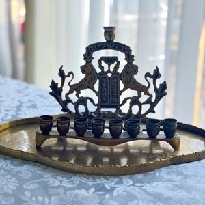 May include: A brass menorah with a decorative lion design and Hebrew text, set on a gold-toned tray. The menorah holds eight small cups for candles. The tray has a hammered texture and curved edges.