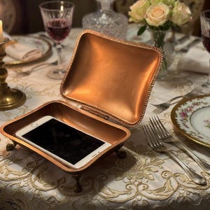 May include: An open, copper-colored, rectangular trinket box with a white smartphone inside, resting on a table. The box has small feet and a hinged lid. The table is set with a lit candle, wine glasses, and floral china.