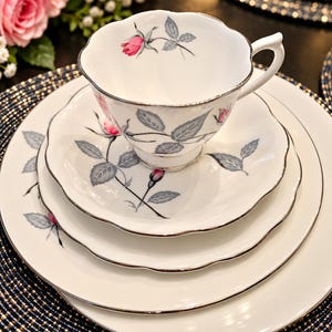 May include: A white porcelain teacup, saucer, and two plates featuring a scalloped edge and a silver rim. The set is decorated with a pattern of pink rosebuds and grey leaves. The teacup has a curved handle and a footed base.