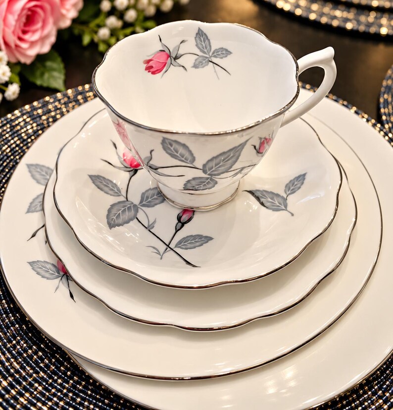 May include: A white porcelain teacup, saucer, and two plates featuring a scalloped edge and a silver rim. The set is decorated with a pattern of pink rosebuds and grey leaves. The teacup has a curved handle and a footed base.