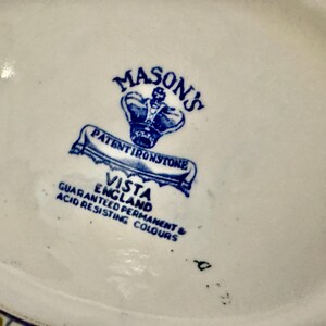 Op de afbeelding: De onderkant van een keramisch object bevat een blauwe stempel met een kroonicoon en de tekst MASON'S PATENT IRONSTONE VISTA ENGLAND GUARANTEED PERMANENT & ACID RESISTING COLOURS. De basis is gebroken wit met een gladde, geglazuurde afwerking.