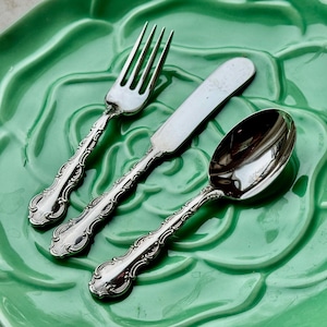 May include: A silver fork, butter knife, and spoon sit on a green, floral-shaped plate. The cutlery has ornate, detailed handles. The plate's surface has a glossy finish. This set is ideal for serving a meal.