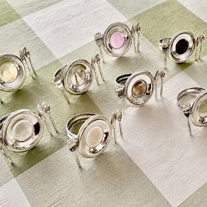 Op de afbeelding: Een set van acht zilverkleurige metalen servetringen, elk voorzien van een miniatuurbordontwerp geflankeerd door een kleine vork en lepel. Het midden van elke ring bevat een cirkelvormige inleg in diverse kleuren, waaronder roze, zwart en wit.