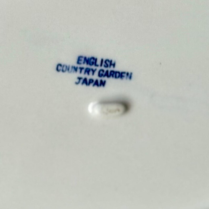 May include: Close-up of a white ceramic surface with blue text that reads "ENGLISH COUNTRY GARDEN JAPAN". A small, oval-shaped mark is below the text. The image suggests a piece of tableware.